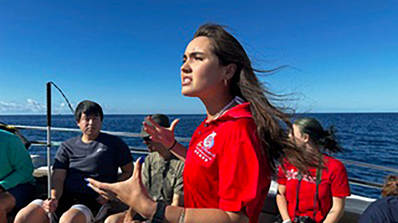 Volunteers gathered on a boat with the ocean in the background. A woman in a red shirt stands at the front, speaking passionately to the group, with a clear blue sky above.