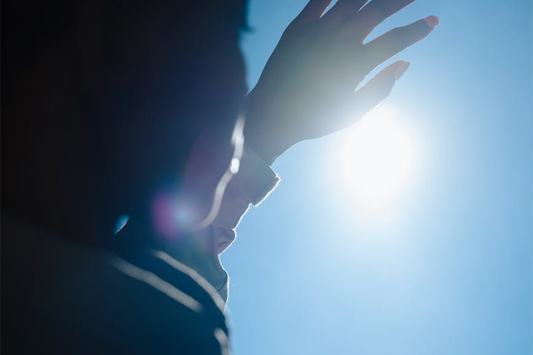 A silhouetted person raises their hand to shield their face from a bright sun in a clear blue sky. The sunlight creates a soft lens flare effect, highlighting the outline of the hand and casting shadows on the person's face.