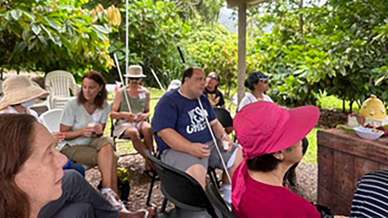 Volunteers gathered outdoors at Foster Botanical Gardens, smiling and holding a sign for an audio scavenger hunt, surrounded by lush greenery.