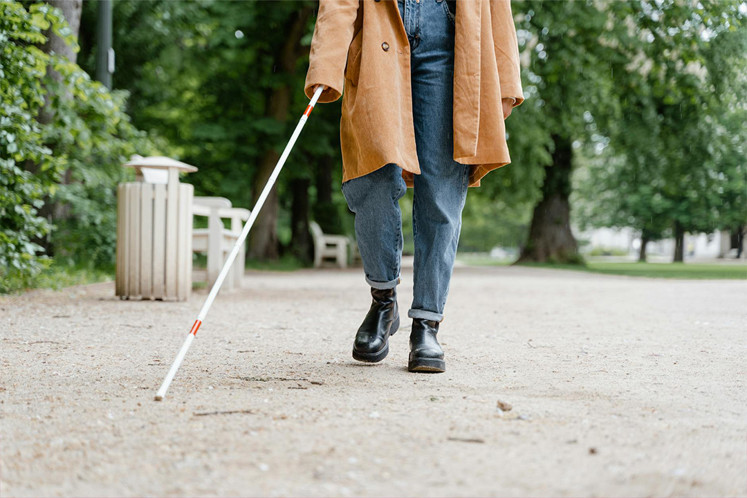 A person walking on a gravel path in a park, holding a white cane with red markings to assist with navigation. They are dressed in a brown coat, jeans, and black boots, with trees and benches visible in the background, conveying a sense of independence and mobility.