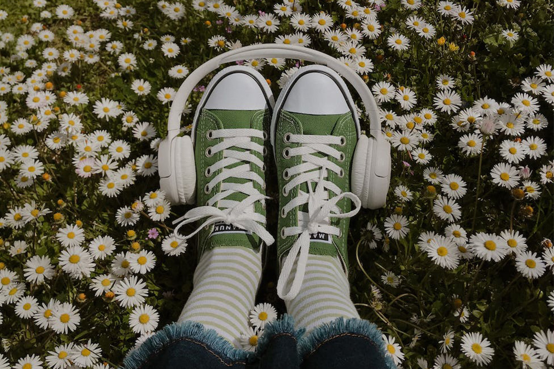 A pair of green sneakers with white laces and a pair of over-ear headphones placed around them, lying in a field of small white daisies. The person wearing the shoes has rolled-up jeans and striped green-and-white socks, creating a relaxed, playful scene.