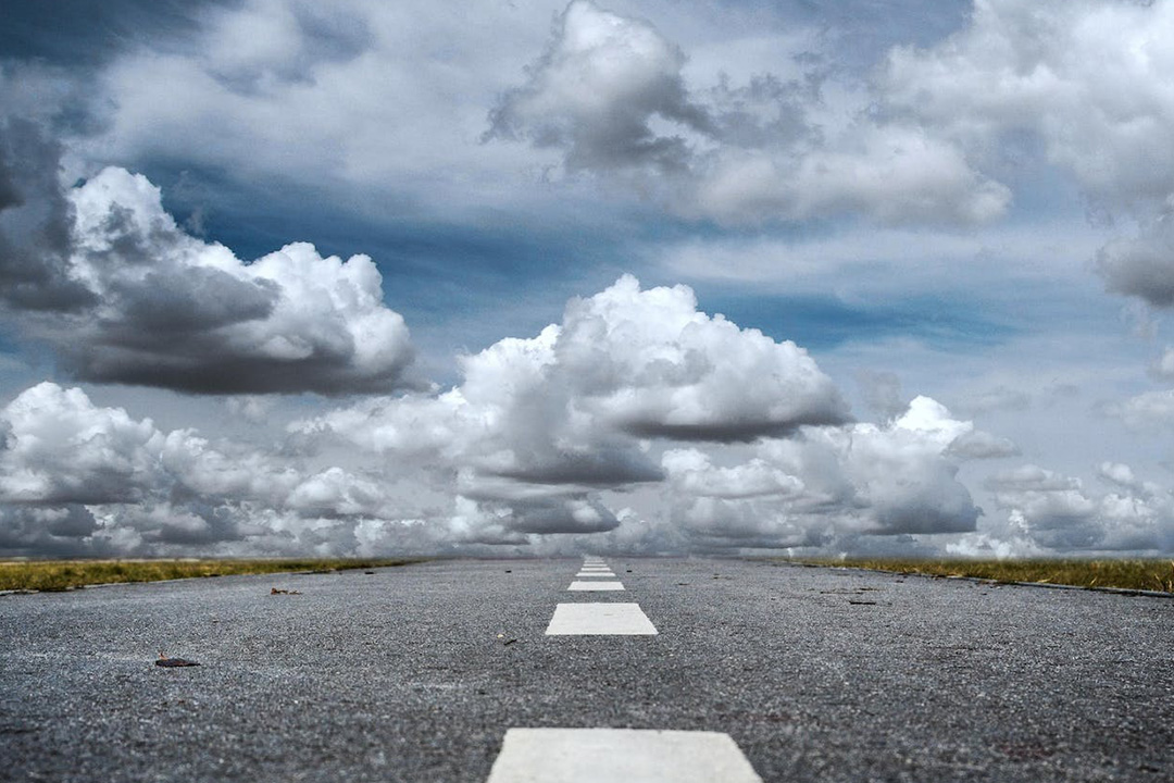 A road extending into the horizon, marked by a white dashed center line. The road is flanked by fields of grass under a vast, cloudy sky with large, billowy clouds casting shadows. The perspective gives the impression of a long, open journey ahead.