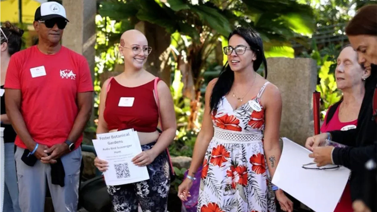 Volunteers gathered outdoors at Foster Botanical Gardens, smiling and holding a sign for an audio scavenger hunt, surrounded by lush greenery.